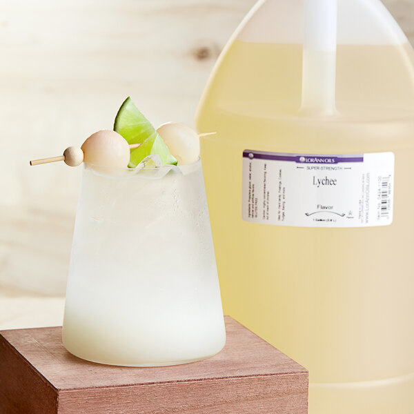 A jug of lychee-flavored liquid on a wooden surface with a glass of lychee-flavored liquid with lime slices.