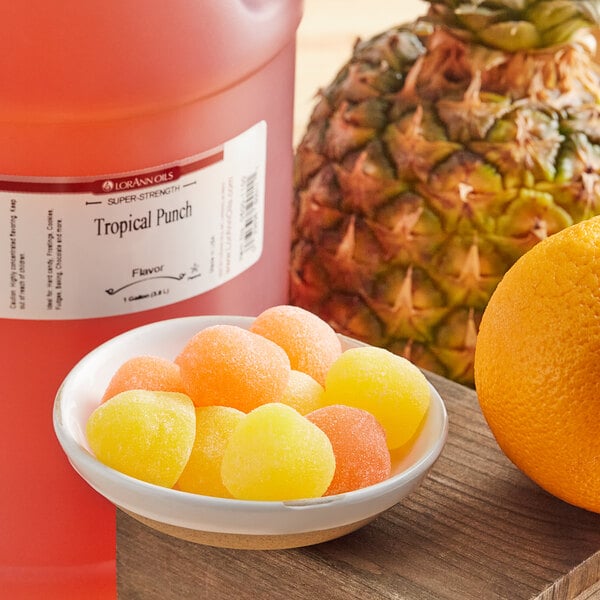 A bowl of gummy candies next to a bowl of yellow and orange candy with a pineapple and a bottle of punch.