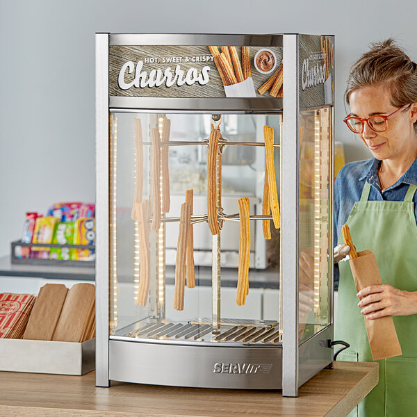 A woman wearing a green apron uses a ServIt countertop display warmer to hold a brown bag of churros.
