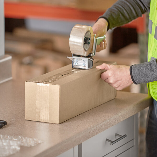 A person wearing a safety vest and using a tape dispenser to seal a Lavex Kraft corrugated shipping box.