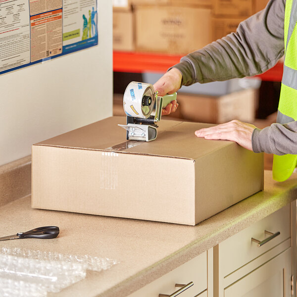 A person using a tape dispenser to seal a Lavex Kraft corrugated shipping box.
