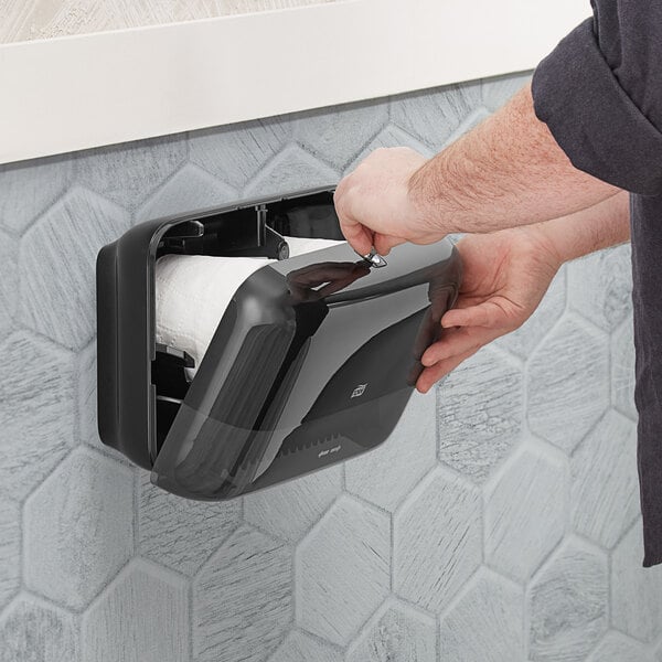 A person opening a black Tork Elevation toilet paper dispenser.