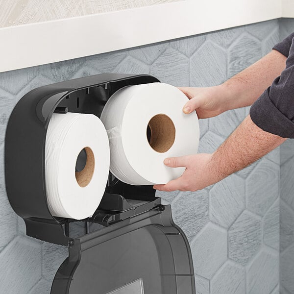 A person putting a Tork Universal jumbo toilet paper roll into a black toilet paper holder.