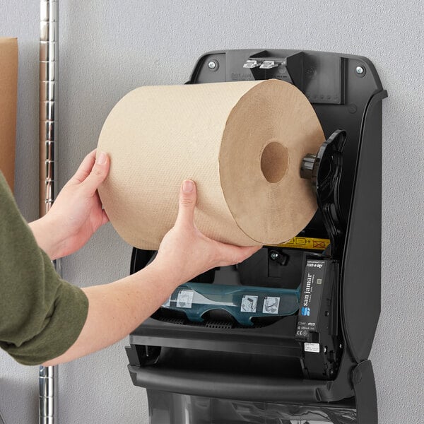 A person holding a Tork Natural Kraft paper towel roll in front of a paper towel dispenser.