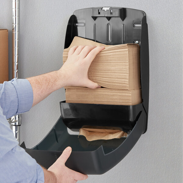 A person putting a stack of Tork natural brown paper towels into a wall-mounted paper towel dispenser.