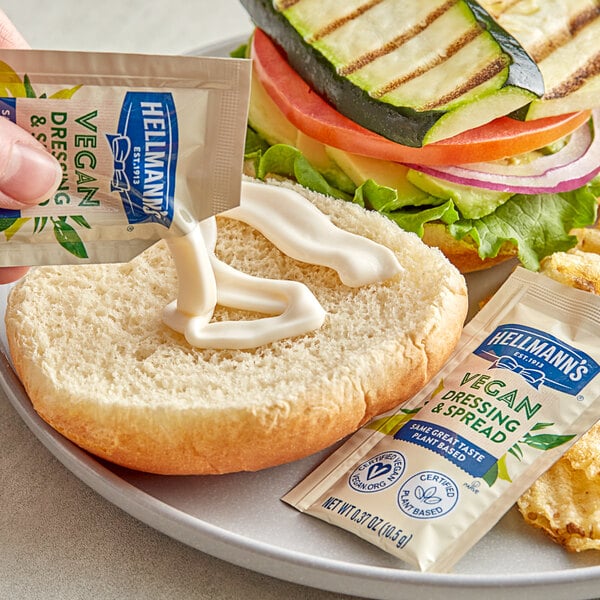 A person pouring Hellmann's vegan mayonnaise from a packet onto a sandwich with vegetables.