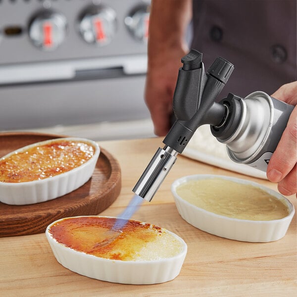 A person using a Chef Master Cooking Butane Torch to cook food over a white dish.