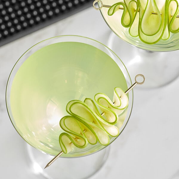 A glass with a drink and a Barfly gold cocktail pick with a green apple spiral on it.