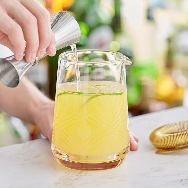 A person pouring yellow liquid into a Barfly stirring glass with lime slices.