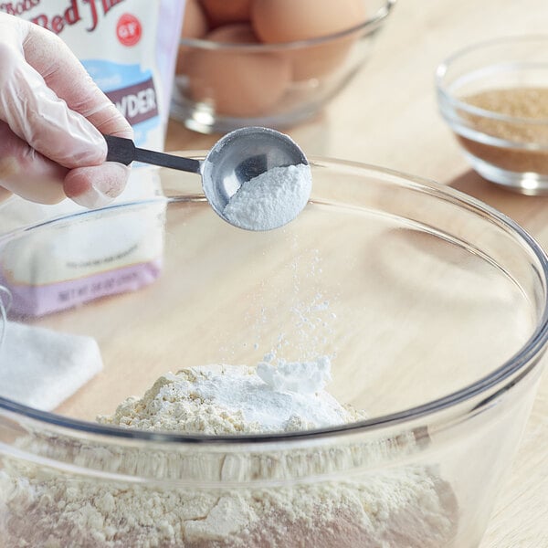A gloved hand mixes Bob's Red Mill gluten-free double-acting baking powder into flour in a bowl with a spoon.