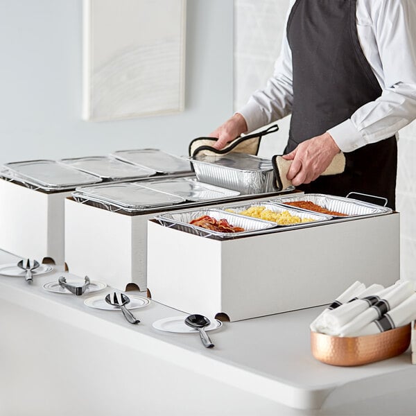 A man in an apron standing next to a hotel buffet with trays of food on table using Choice chafing dishes.