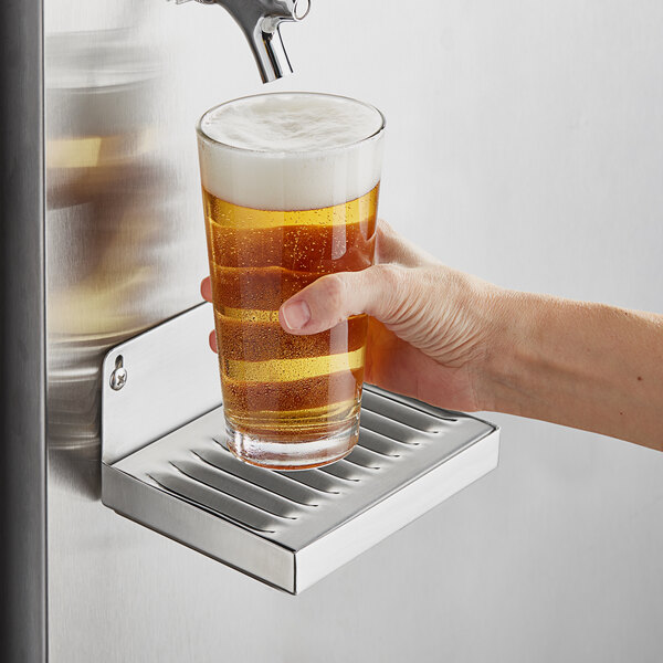 A hand holding a glass of beer under a Regency stainless steel wall mount beer drip tray.