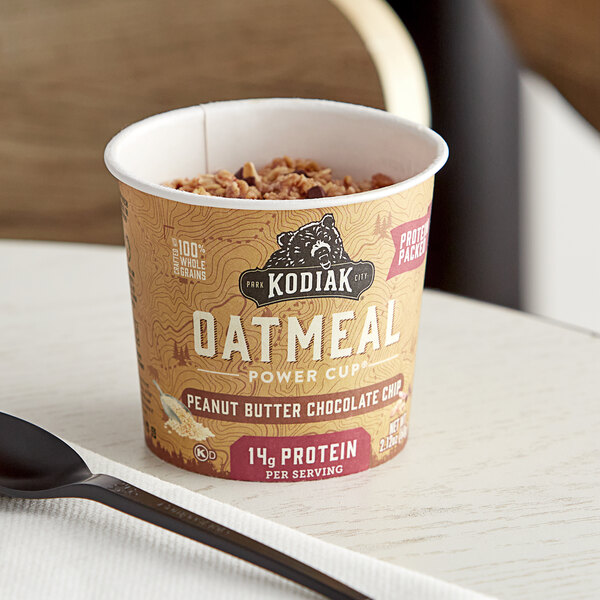 A close-up of a Kodiak Cakes Peanut Butter Chocolate Chip Oatmeal Cup on a table with a bowl of oatmeal.