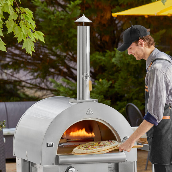A man using a wooden peel to cook a pizza in a Backyard Pro stainless steel wood fired pizza oven.