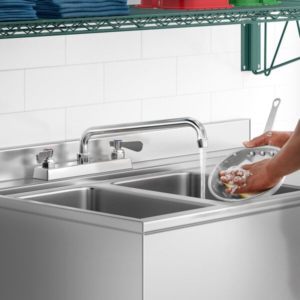 A person using a Regency deck-mounted faucet to wash dishes in a stainless steel sink.