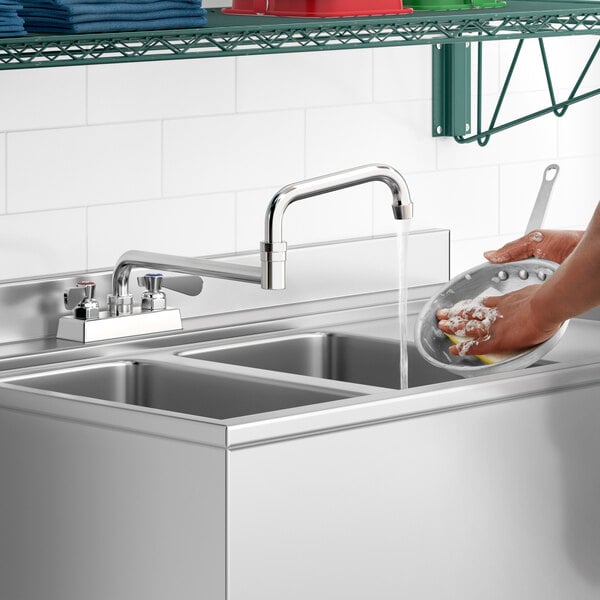 A person using a Regency deck-mounted faucet with a double-jointed swing spout to wash dishes in a stainless steel sink.