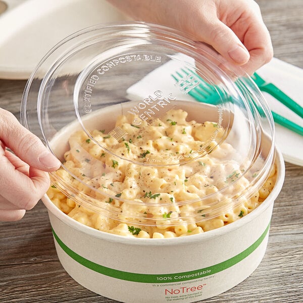 A hand holding a World Centric clear plastic lid over a bowl of macaroni and cheese.