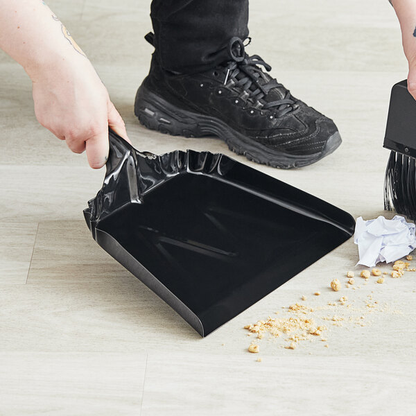 A person using a Lavex black steel dust pan to sweep the floor.