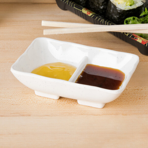 A white rectangular melamine sauce dish with 2 sections holding a couple of different types of sauces.