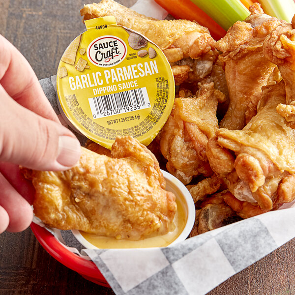 A hand holding a Sauce Craft Garlic Parmesan dipping sauce container with a bowl of chicken wings.
