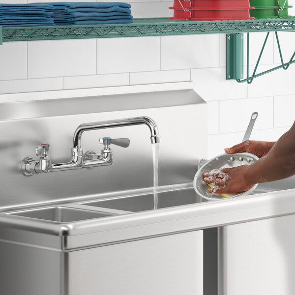 A person using a Regency wall mount faucet with a swing spout to wash dishes in a stainless steel sink.
