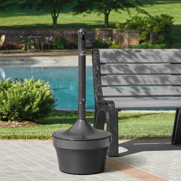 A black Rubbermaid Aladdin cigarette receptacle on a metal pole by a bench in a park.