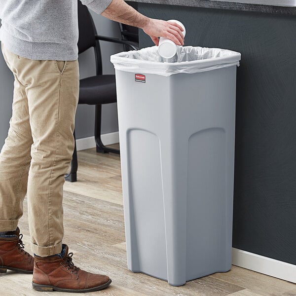 A man putting a cup into a Rubbermaid Untouchable 23 gallon gray square trash can.