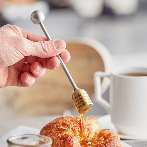 A hand holding an Acopa stainless steel honey dipper over a croissant.