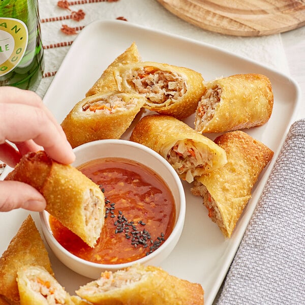 A hand holding a fried vegetable egg roll with a plate of egg rolls and dipping sauce.