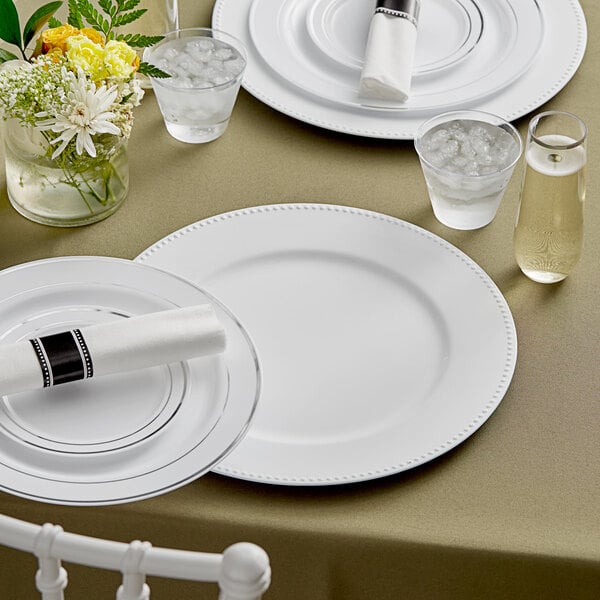 A table set with white Choice beaded rim charger plates, silverware, and flowers.
