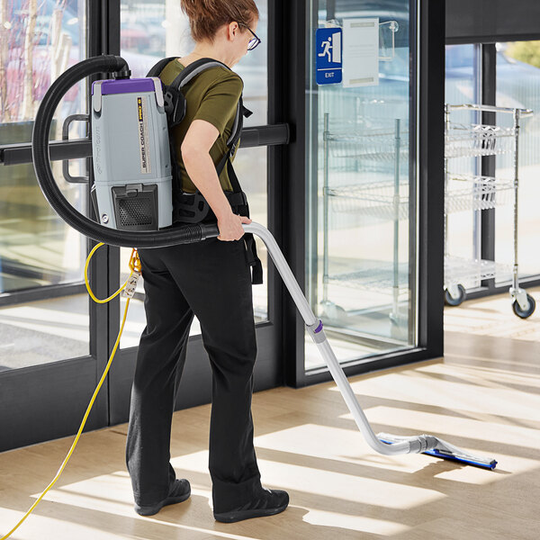 A woman using a ProTeam backpack vacuum cleaner in a large room.