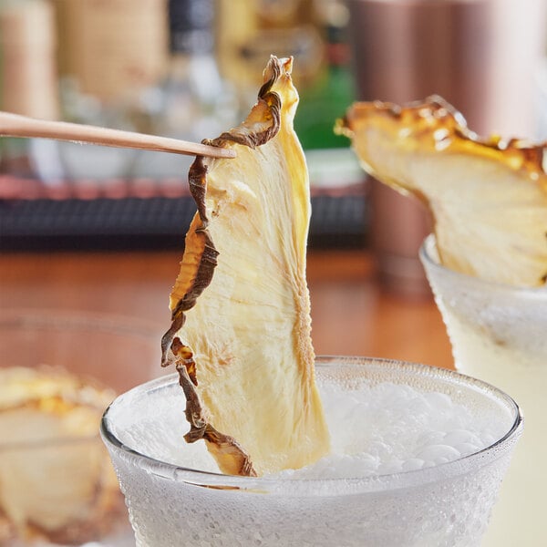 A dried pineapple half slice on a stick being placed in a glass of ice.