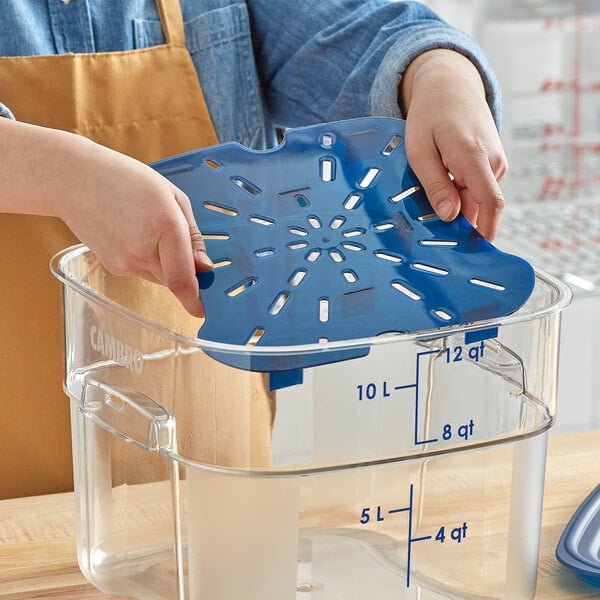 A woman using a measuring cup to fill a blue Cambro food storage container with food.