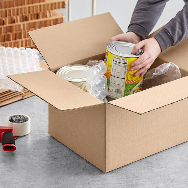 A person packing a Lavex heavy-duty corrugated cardboard shipping box with cans and other items.