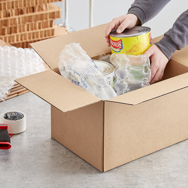 A person holding a can of food and putting it in a Lavex Kraft cardboard shipping box.