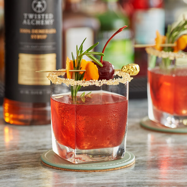 A glass of red liquid with a garnish of fruit and rosemary on a table with a bottle of Twisted Alchemy Demerara Syrup.