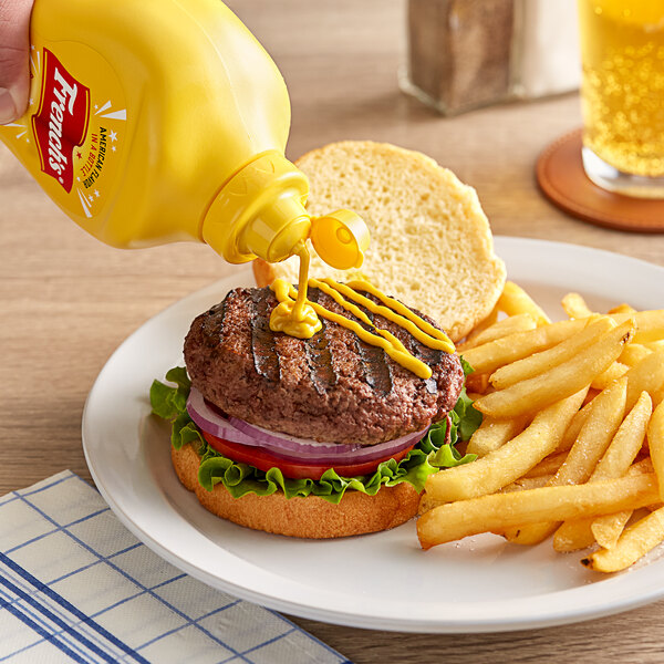 A hand pouring French's Classic Yellow Mustard onto a burger and fries on a plate.