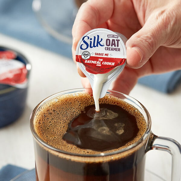 A hand pouring Silk oat milk creamer into a cup of coffee.