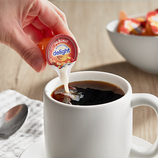 A hand pouring International Delight Hazelnut creamer into a cup of coffee.