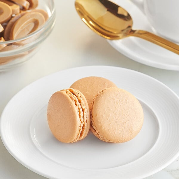 A white plate with a brown macaron, a bowl of brown macarons, and a gold spoon.