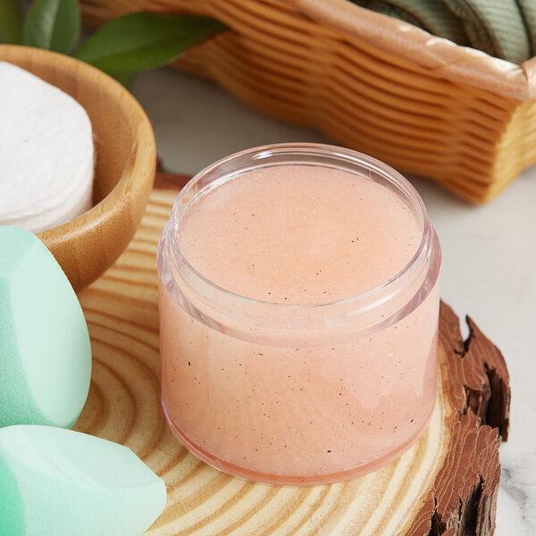 A 4 oz. clear plastic jar of pink face scrub on a wood surface next to a basket of sponges.