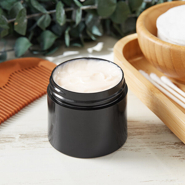 A black container of white cream next to a wooden bowl with a comb and cotton swabs.