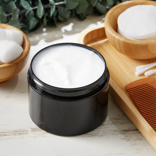 An 8 oz. black plastic jar of white cream next to a wooden bowl of cotton pads and a wooden comb.