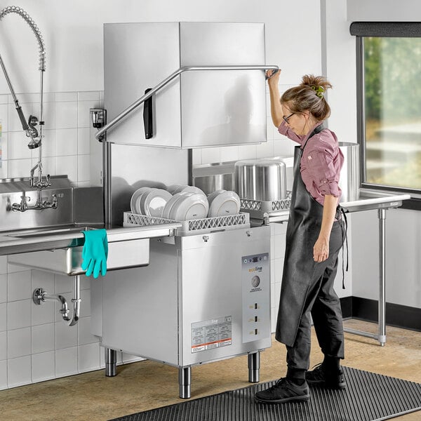 A woman in overalls cleaning an Ecoline by Hobart door-type dishwasher on a counter in a professional kitchen.