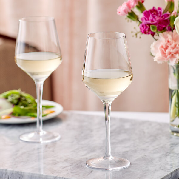 Two Acopa Silhouette wine glasses filled with white wine on a marble table with flowers.