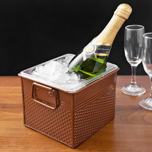 A bottle of champagne in a hammered copper stainless steel square beverage bucket with ice and two wine glasses.