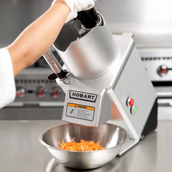 A person using a Hobart food processor to cut carrots into a bowl.