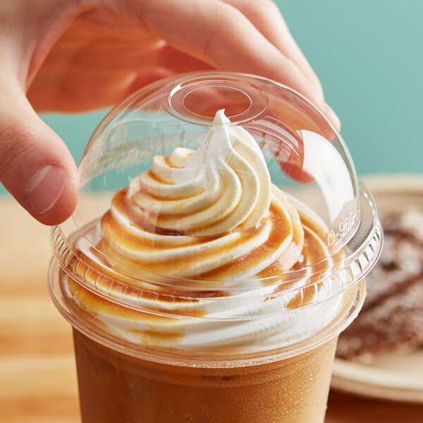 A hand holding a New Roots compostable plastic cup with a whipped cream topping and a dome lid with an opening.