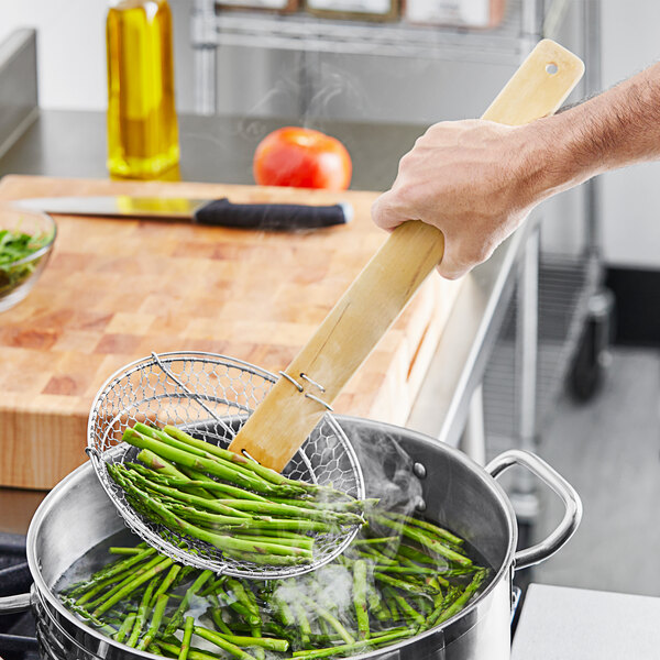 A person using Emperor's Select Coarse Skimmer with a bamboo handle to cook asparagus in a frying pan.