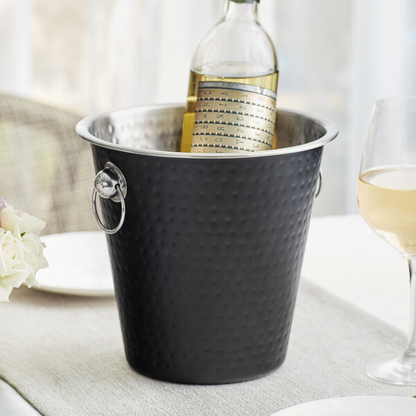 A wine bottle in a black Acopa stainless steel wine bucket on a table on an outdoor patio.